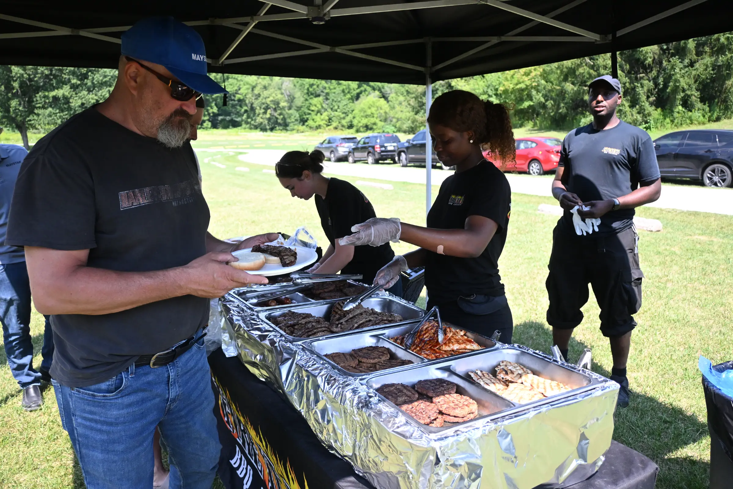 A man wearing a blue cap and sunglasses receives food at an outdoor buffet setup under a tent, served by catering staff.