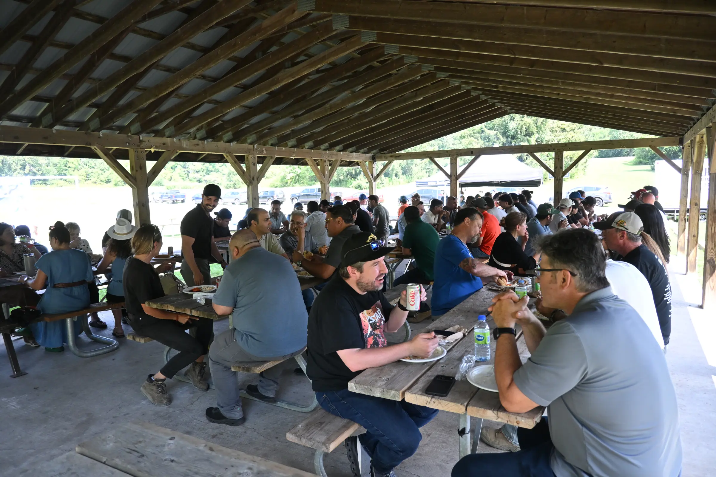 Maystar team members eating lunch and chatting together at wooden picnic tables under a shaded park pavilion.